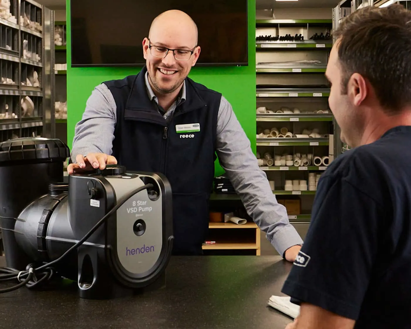 Store employee in blue vest showing a Henden VSD pump to customer, with plumbing supplies on shelves behind them.