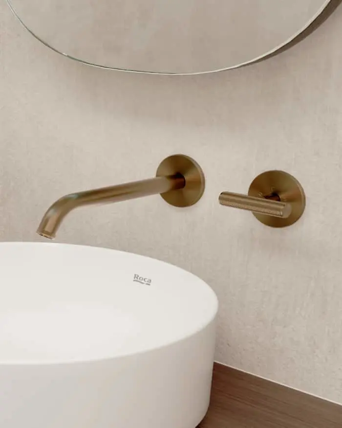 Modern bathroom with brass wall-mounted faucet, white Roca sink, and partial view of a round mirror above.