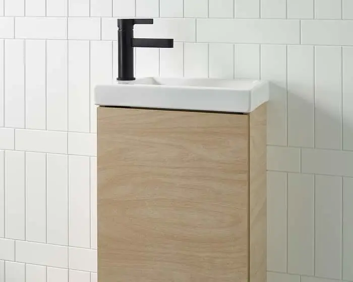 Small bathroom vanity with white ceramic sink, black faucet, and light wood cabinet against white tiled wall.