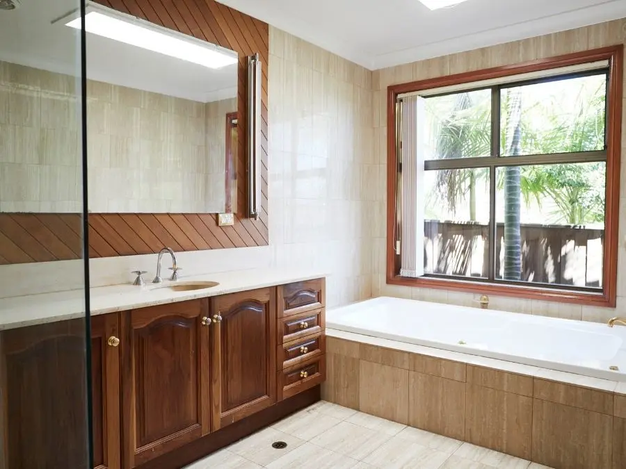 Bathroom with wooden vanity, large mirror, white bathtub, and window overlooking palm trees.