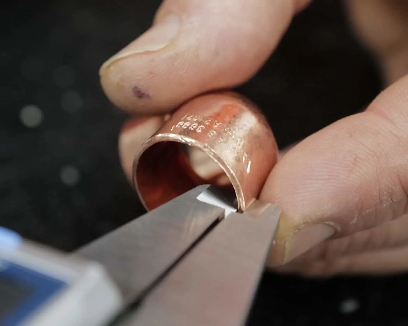 Close-up of hands using pliers to work on a copper ring with engraved numbers against a dark background.