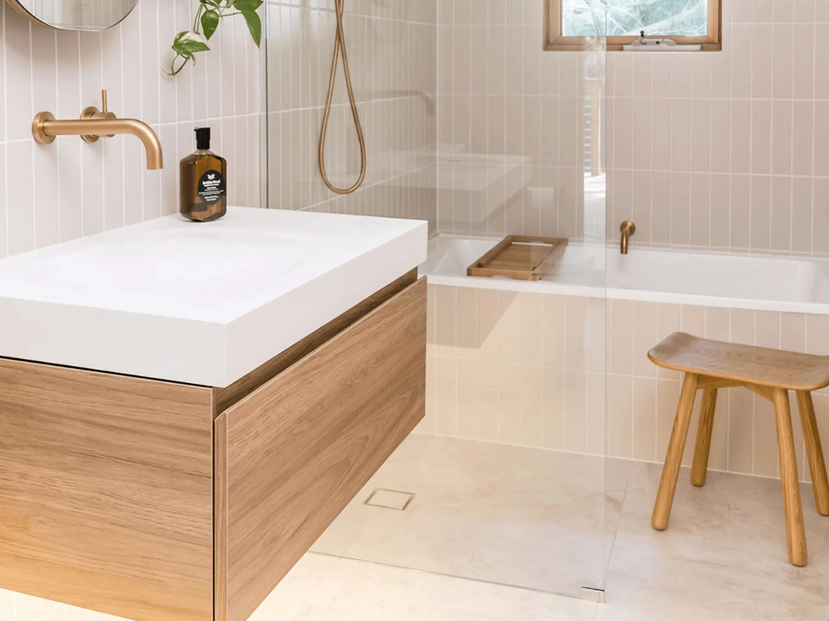 Modern bathroom with white sink, wooden vanity, glass shower, bathtub, and wooden stool. Brass fixtures and cream tiles.