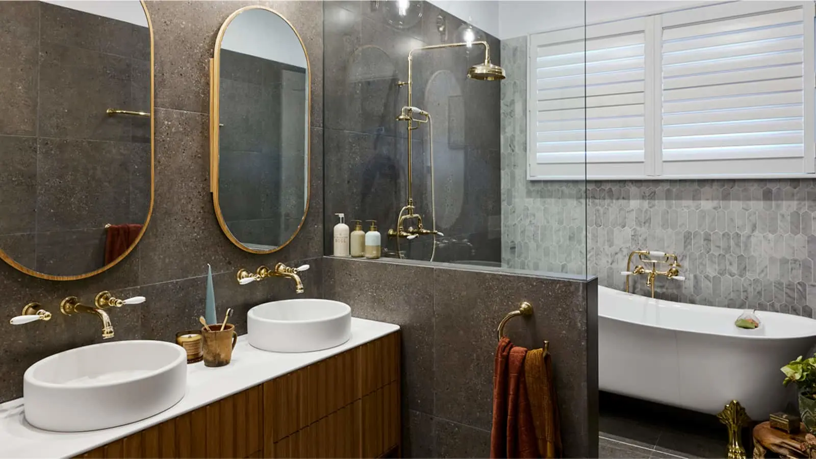 Modern bathroom with dark gray tile, dual white sinks, gold fixtures, oval mirrors, glass shower, and freestanding tub.