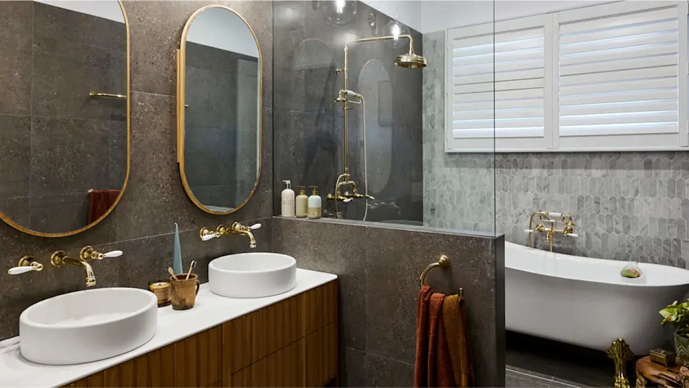 Modern bathroom with dark gray tile, dual white sinks, gold fixtures, oval mirrors, glass shower, and freestanding tub.