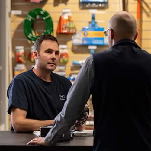 Two people conversing at a hardware store counter with garden supplies displayed on the wall behind them.