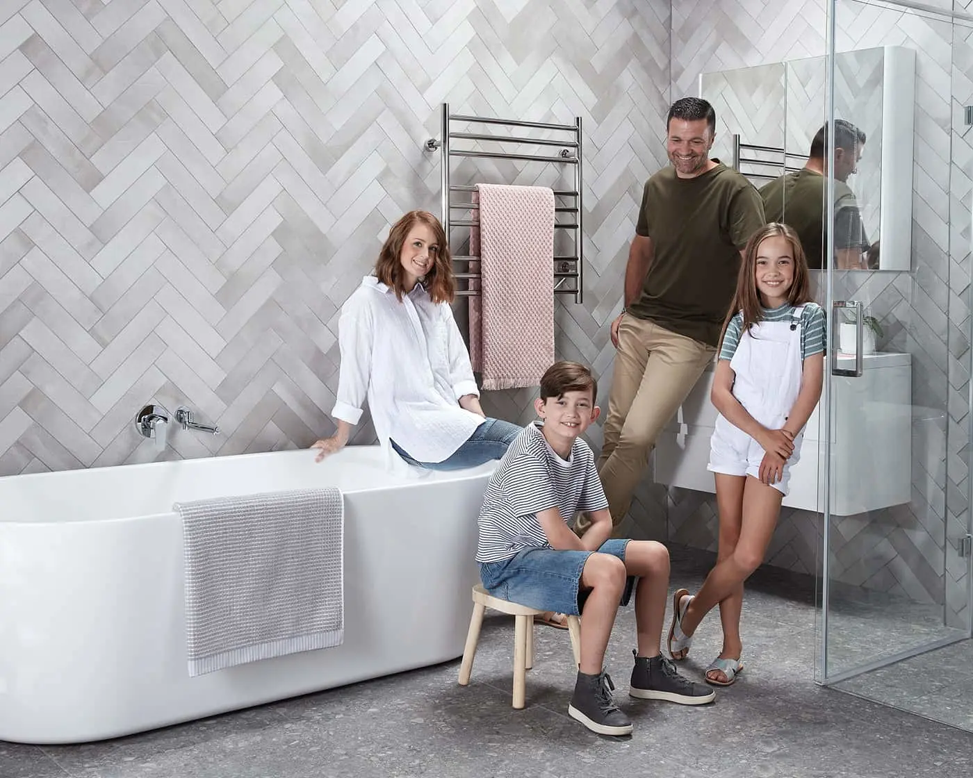 Family in modern bathroom with white herringbone tile walls, freestanding bathtub, heated towel rack, and glass shower enclosure.