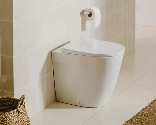 White modern toilet in corner of bathroom with beige tile walls and floor, toilet paper holder mounted on wall.