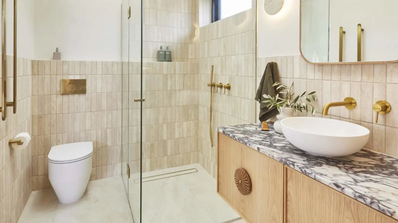 Modern bathroom with beige tiles, glass shower, white toilet, marble countertop with vessel sink, and brass fixtures.