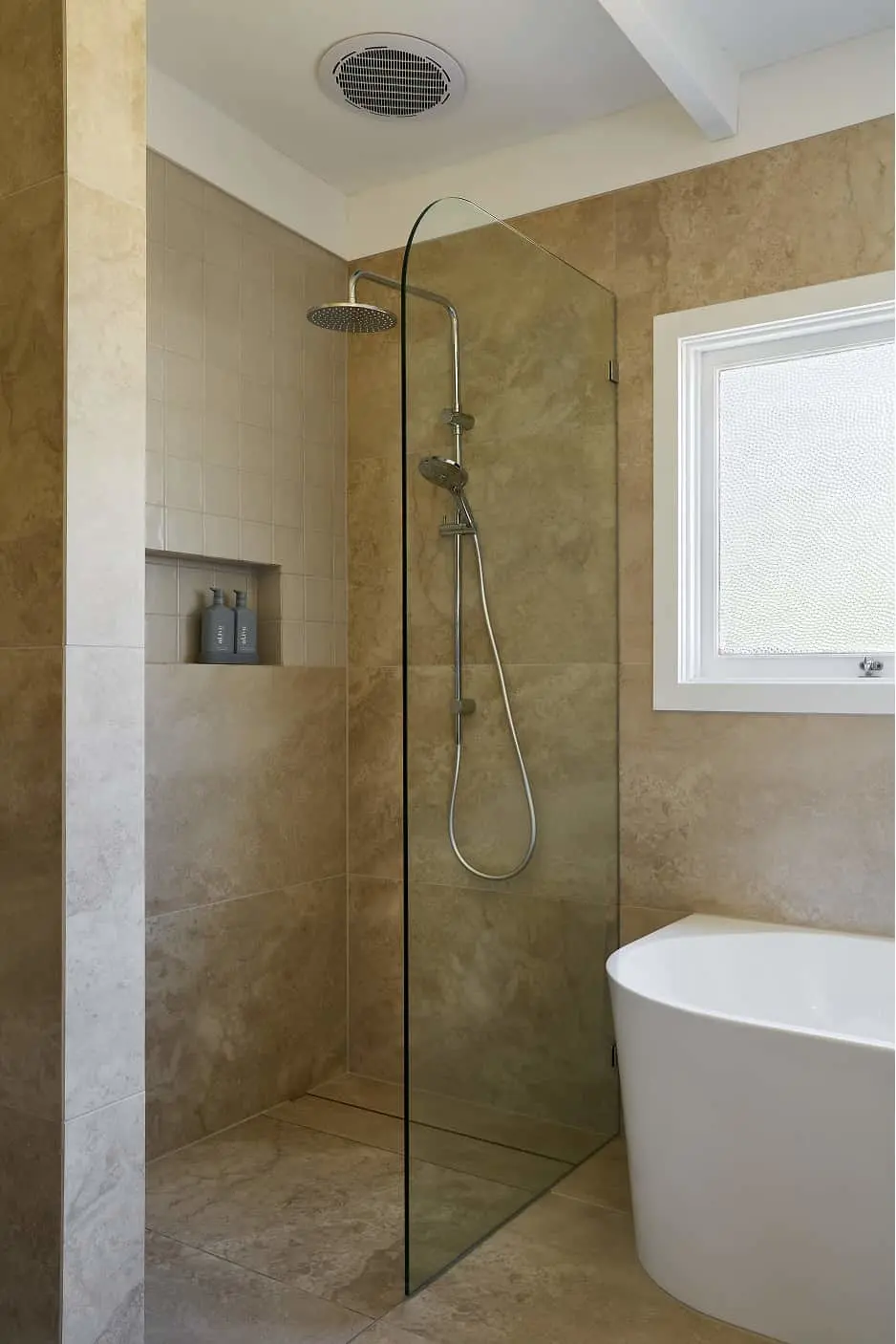 Modern bathroom with walk-in glass shower, beige tile walls, rainfall showerhead, built-in niche, and white bathtub.