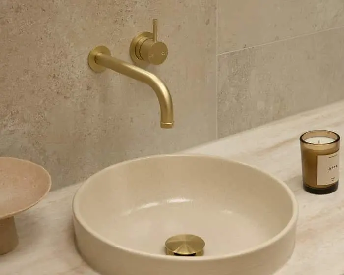 Brass wall-mounted faucet above a cream-colored round vessel sink on a marble countertop with a candle nearby.