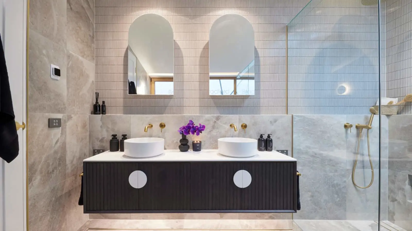 Modern bathroom with dual white vessel sinks on black vanity, marble walls, arched mirrors, and purple flowers.