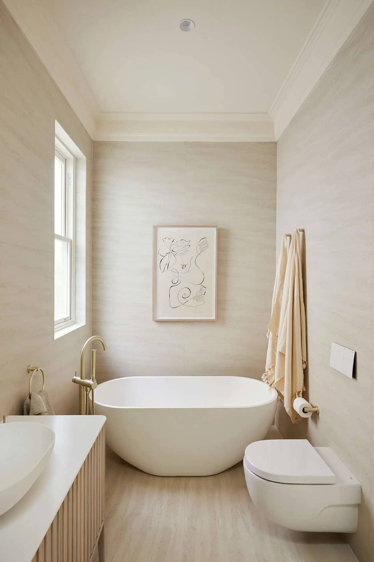 Modern beige bathroom with freestanding white tub, abstract wall art, toilet, and sink with brass fixtures.