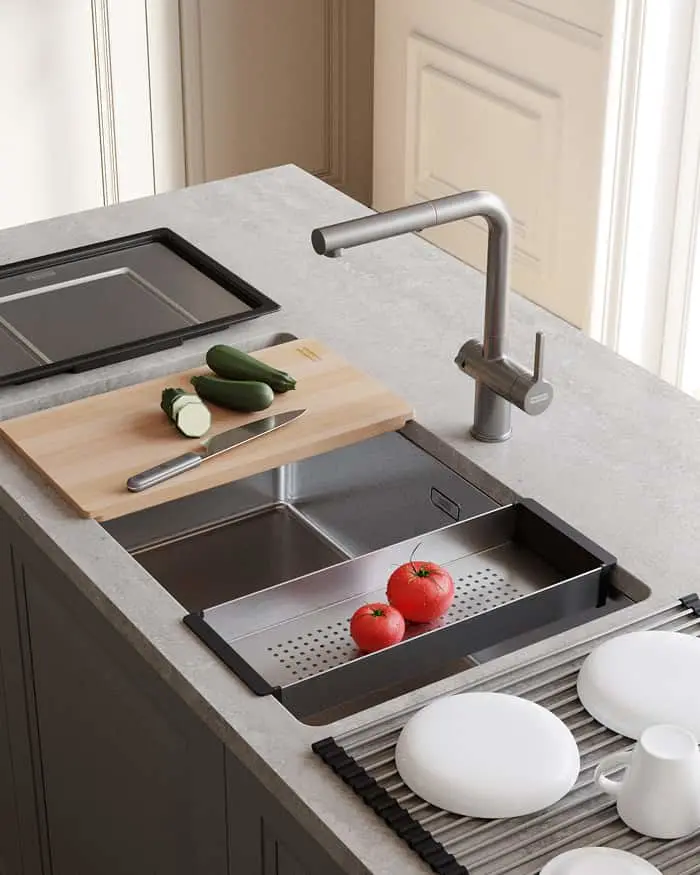 Stainless steel kitchen sink with colander holding tomatoes, wooden cutting board with zucchini, and gray faucet on stone countertop.