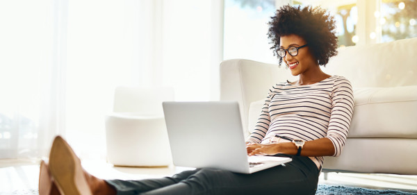 a black woman sits on the floor against a white couch using a laptop