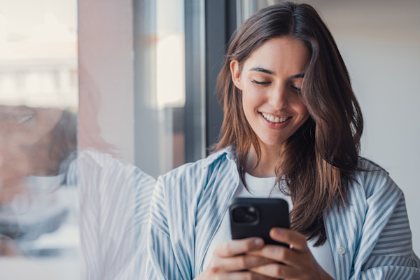 A woman looks at her phone while smiling and standing next to a window