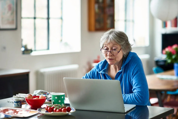 A gray-haired woman in a blue hoodie uses her laptop at a kitchen counter.