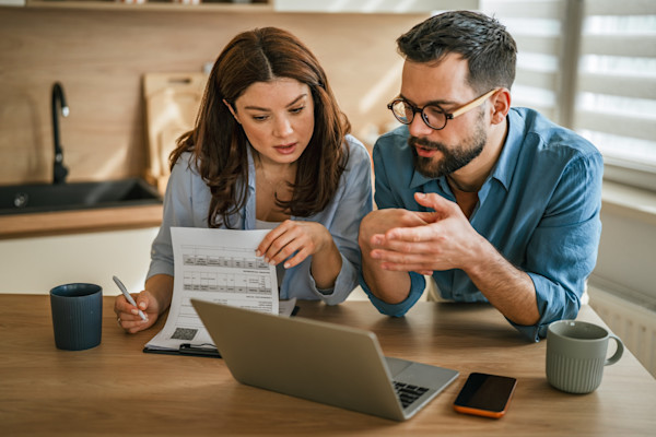 A woman and man discuss bills on a clipboard in front of a computer