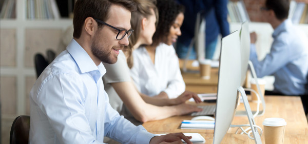 a professional white man in glasses uses a desktop computer with colleagues in the background