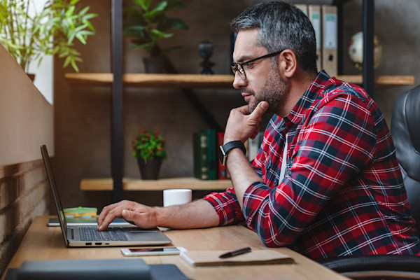 Man with glasses sitting at a desk and looking at his laptop