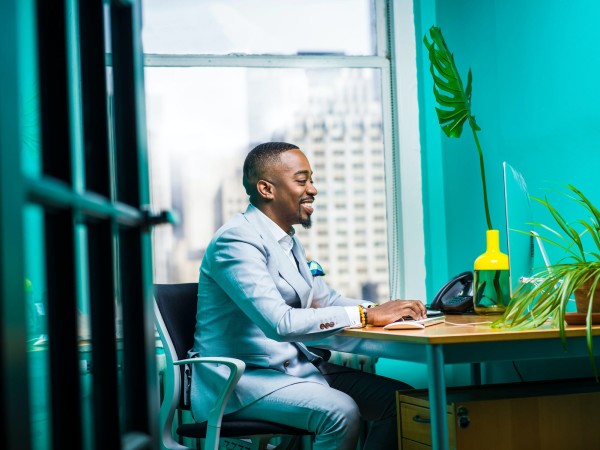 A smiling, professional black man types in his very teal office