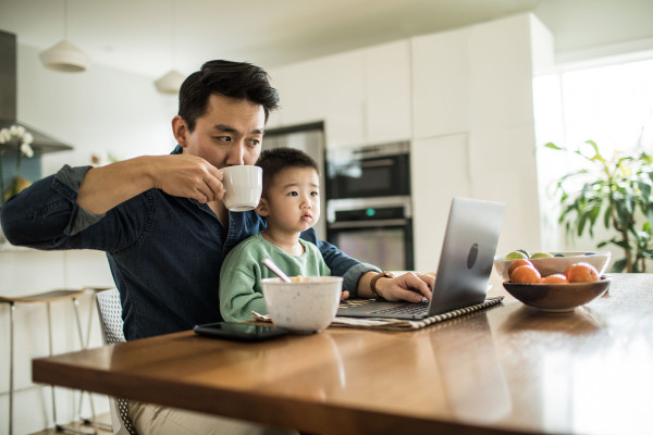 An Asian father and his toddler son sit at the kitchen table looking at a laptop