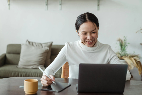 Woman in white sweater sitting at desk writing at laptop with coffee. JPG.