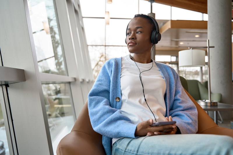 Black woman wearing a blue cardigan looking out the window with headphones on