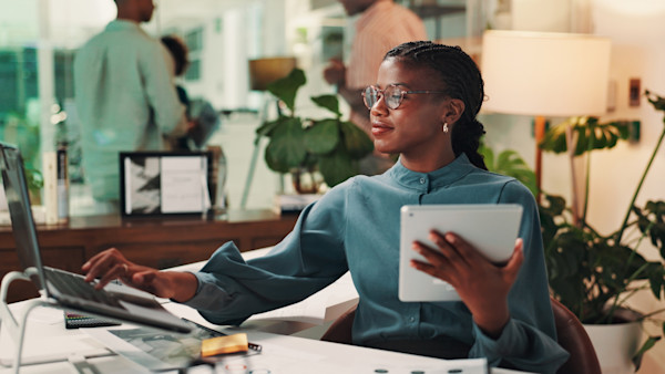 A woman sits at a desk in an office with a computer in front of her and a tablet in one hand