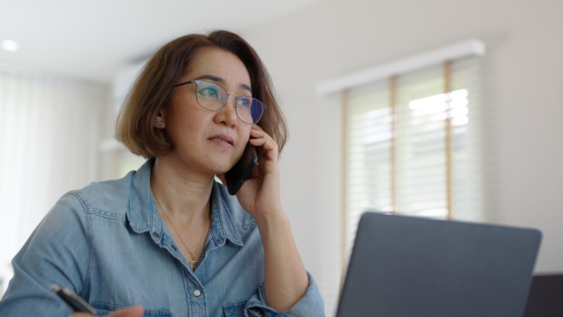 Woman with glasses wearing a denim button down talking on the phone with a laptop in front of her