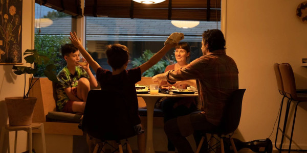 A white-presenting family of four have dinner in their home, and one child sticks his arms up exuberantly.