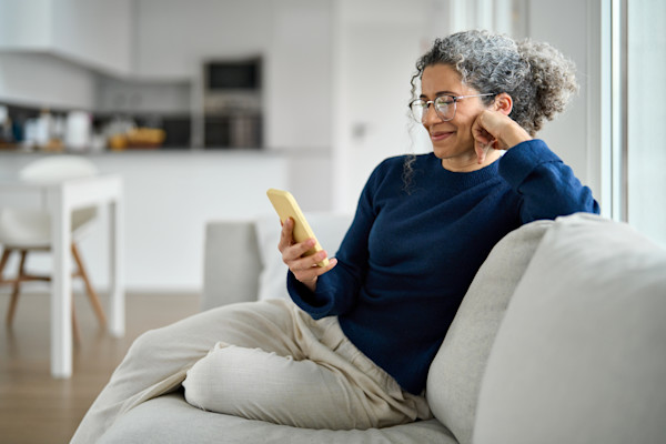 An older woman sits on a couch while scrolling on her phone