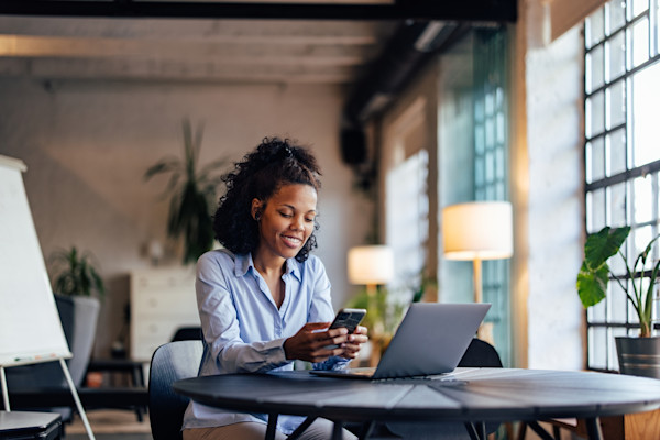 Black woman sitting down at a desk looking at her phone and laptop