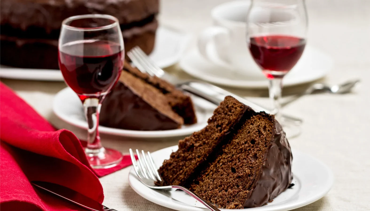 Slices of chocolate cake on plates with glasses of red wine and cutlery set on a light tablecloth.