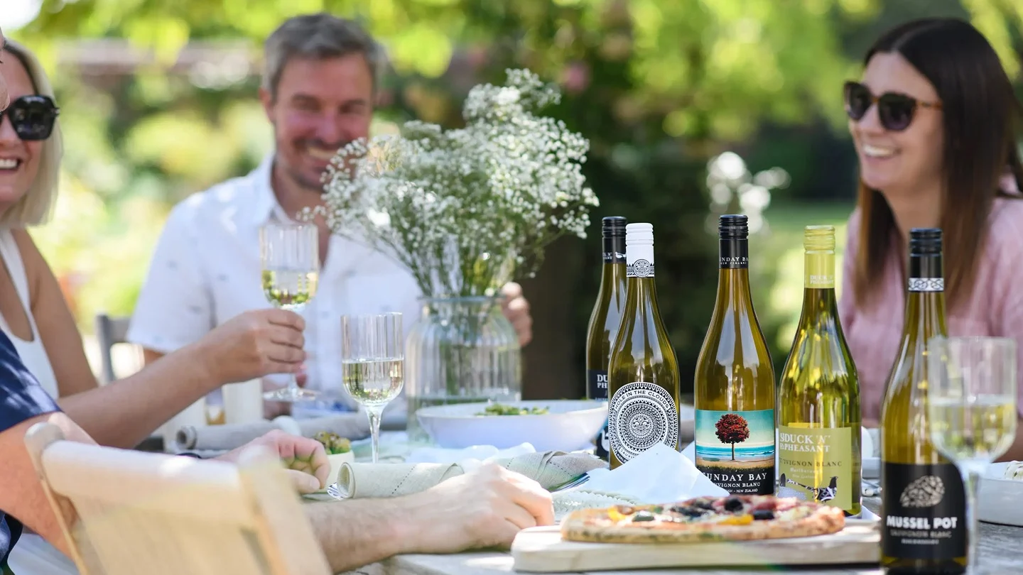 People gathered around an outdoor dining table with bottles and glasses of Sauvignon Blanc wine, food and flowers.
