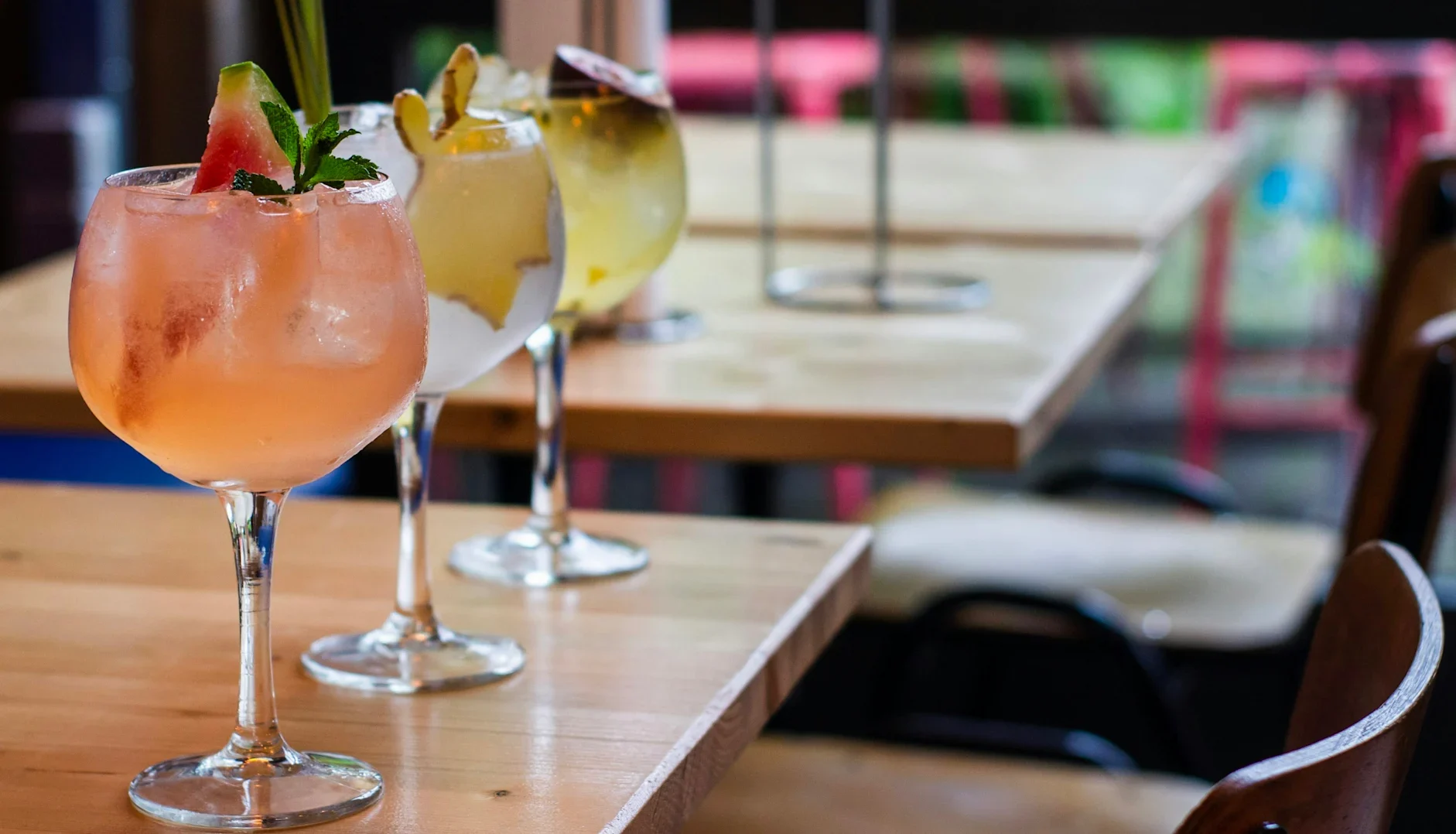 Three colourful cocktails in large stemmed glasses on a wooden table in a bar setting.
