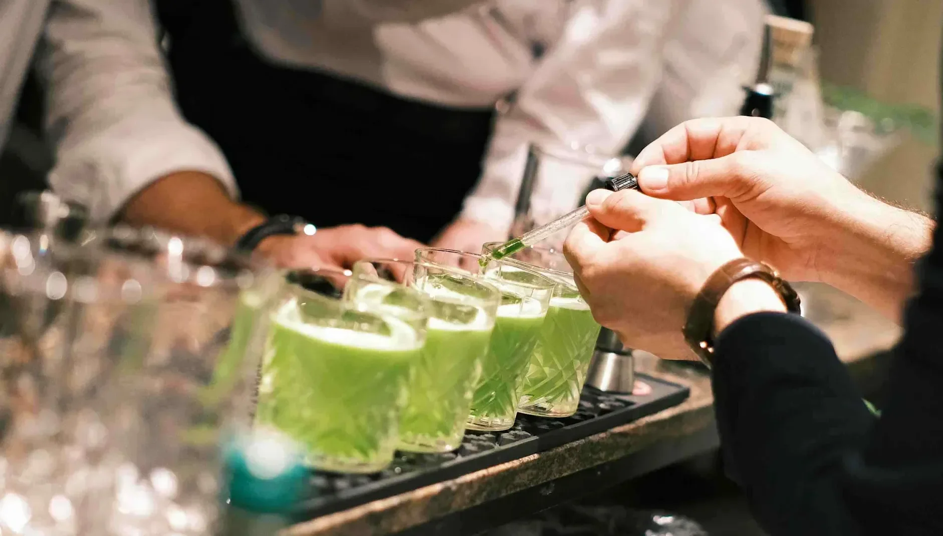 Green cocktails in glasses on a bar counter while a dropper adds garnish during preparation.