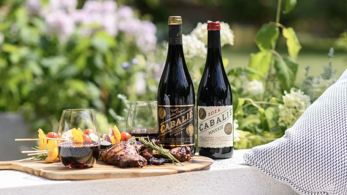Two bottles and glasses of red wine beside grilled meat and skewers on a wooden serving board in a garden setting.