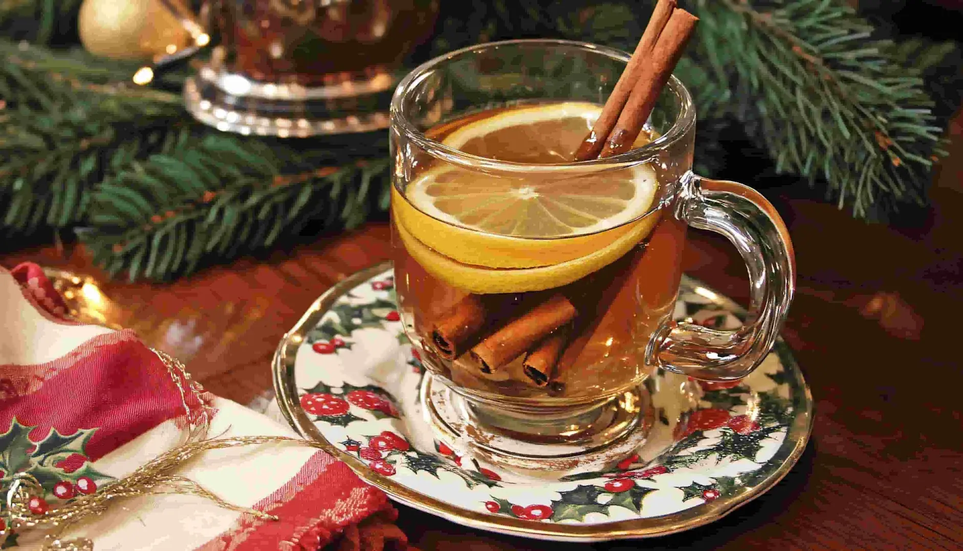 A glass teacup containing a Hot Toddy cocktail is set on a Christmassy table - the teacup contains lemon and cinnamon stick garnish.