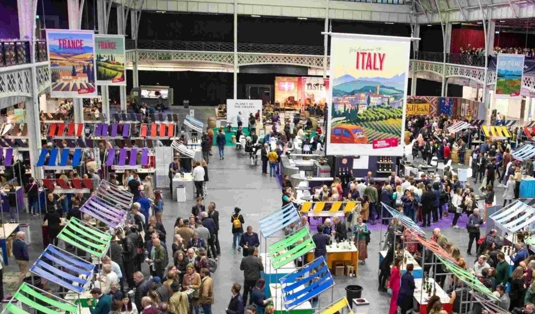 Crowded wine festival hall with country-themed displays and tasting stands beneath a large glass roof.