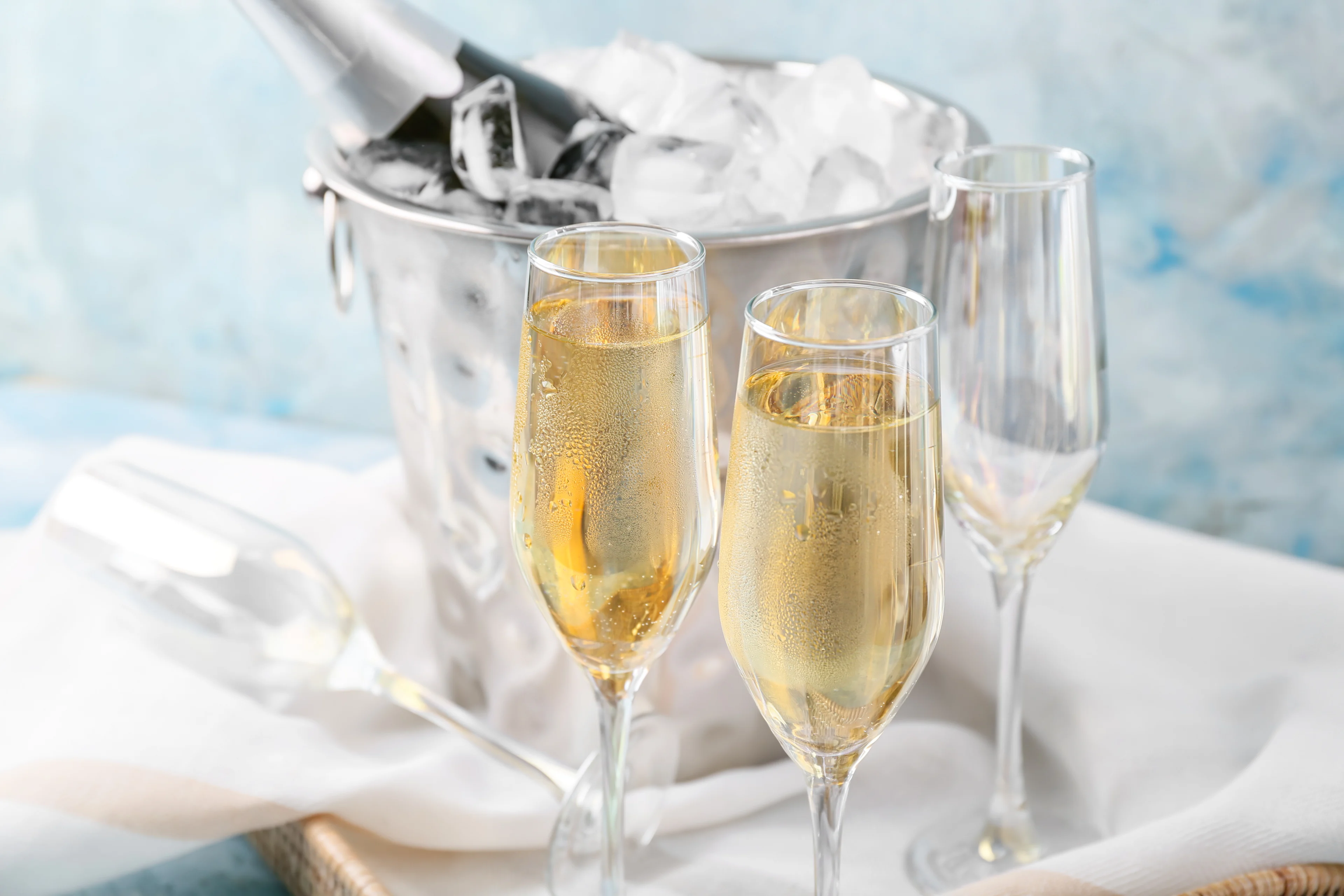 Two filled champagne flutes in front of a silver ice bucket holding a chilled bottle, with an empty glass lying on a white cloth against a light blue background.