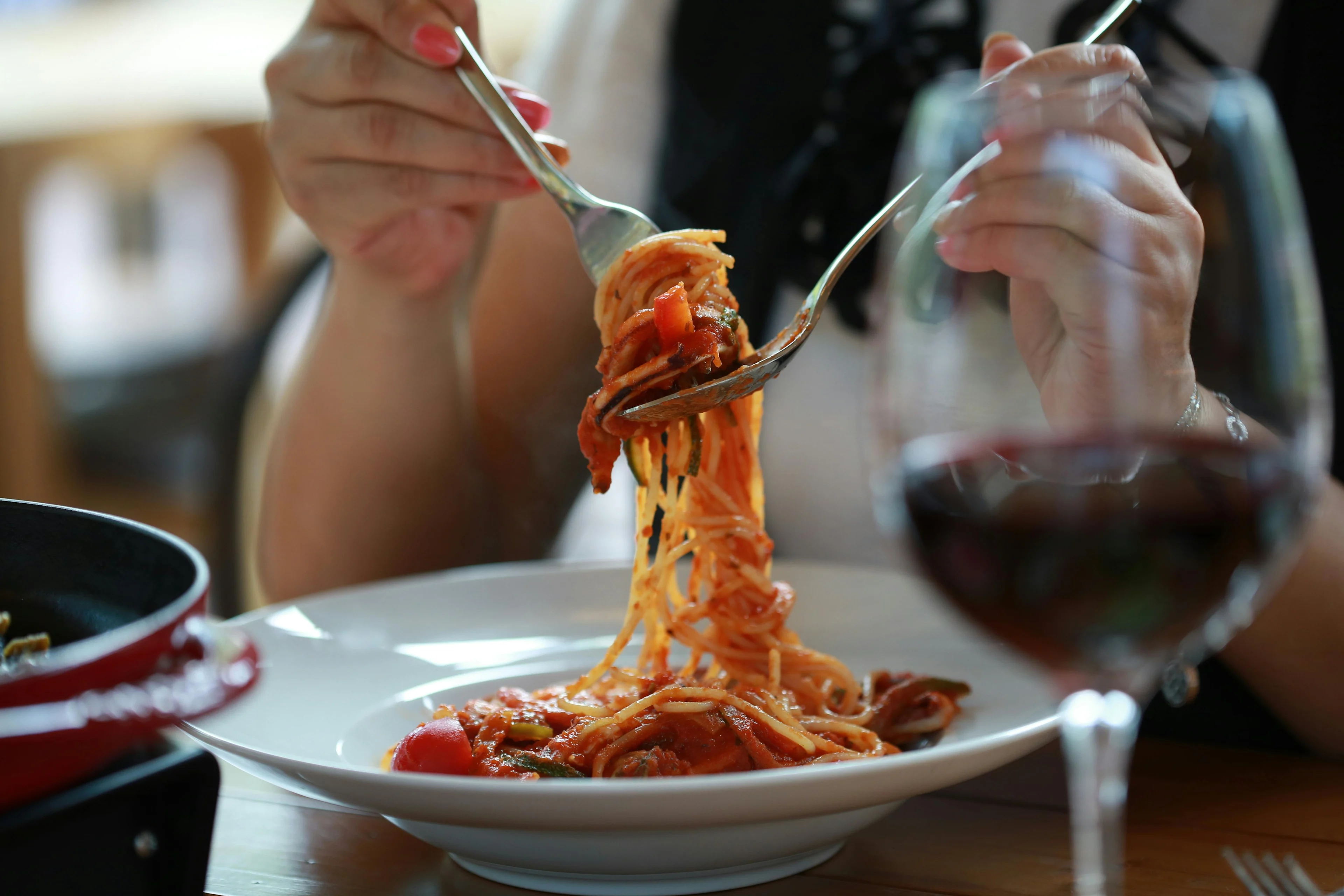 A person twirling spaghetti with tomato sauce on a fork over a white plate, with a glass of red wine in the foreground and a pan of food partially visible on a wooden table.