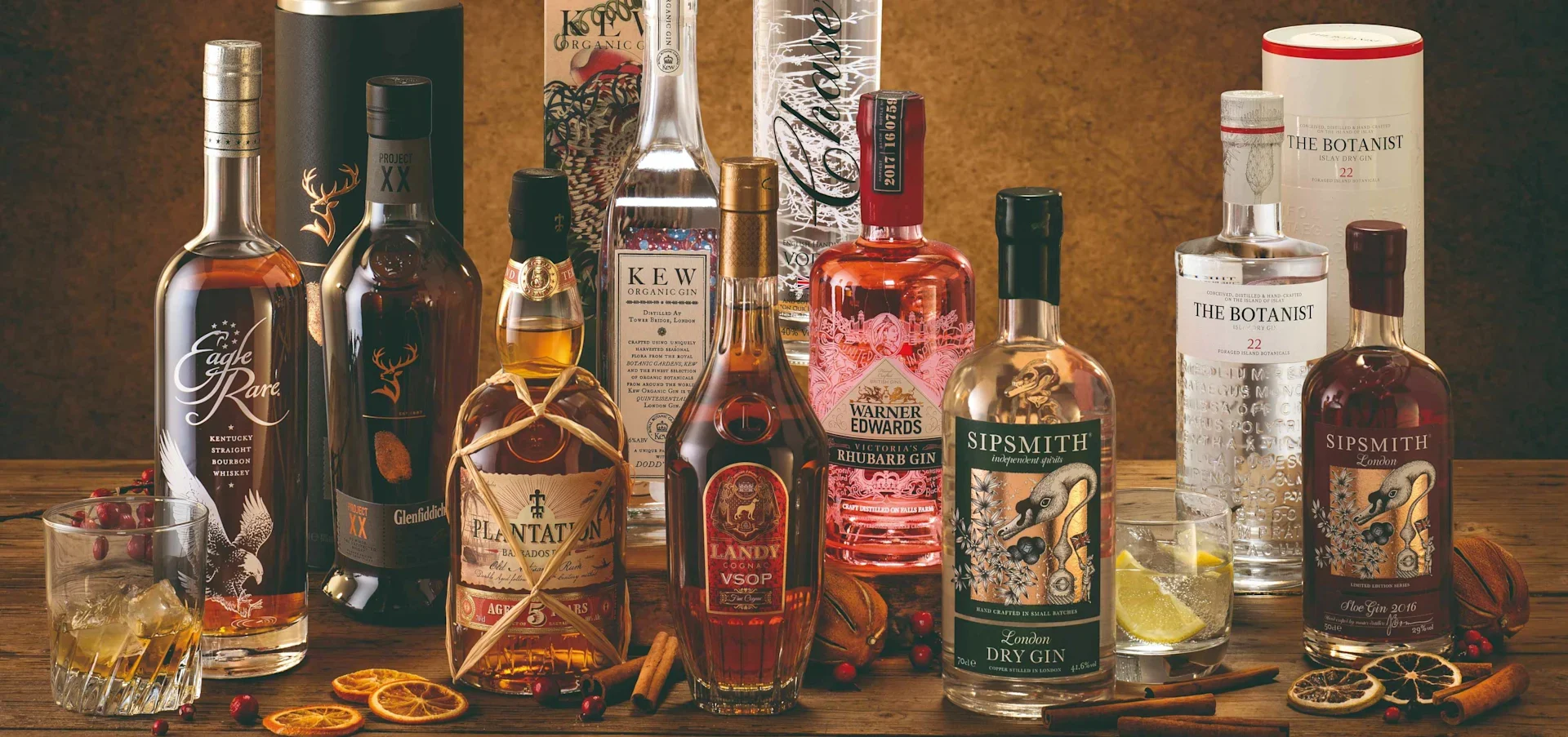Assorted spirits bottles including gin, whiskey, and rum with garnished glasses, dried citrus, and spices on a wooden surface.