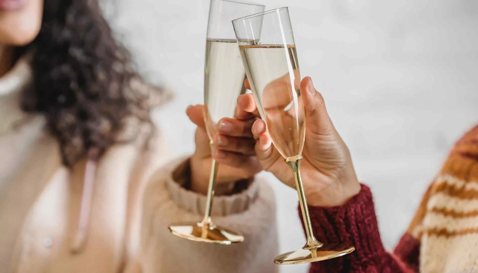 Two people clinking glasses of sparkling wine, each holding a tall flute, with soft indoor lighting and warm sweaters visible.