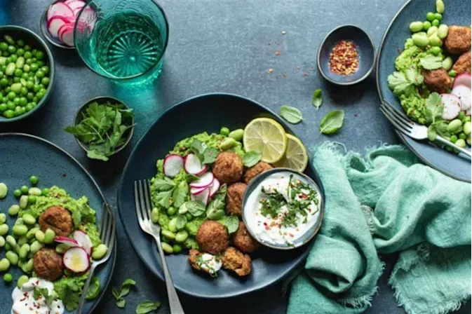 A dark plate with falafel balls, green pea puree, sliced radishes, edamame, fresh herbs, and a bowl of creamy dip, surrounded by additional bowls of greens and spices on a textured surface.