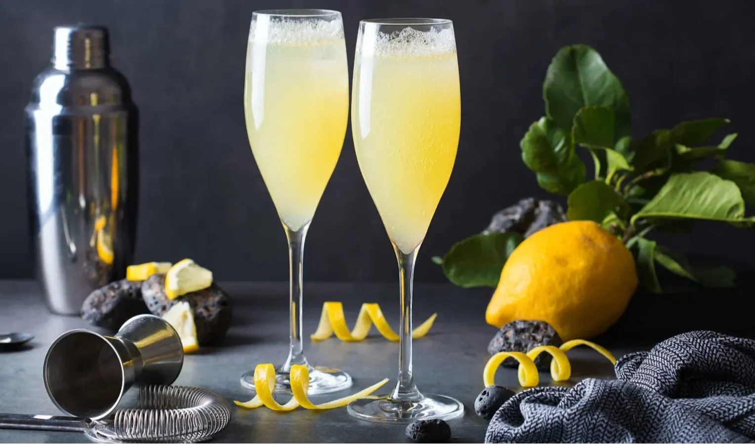 Two tall champagne flutes filled with a pale yellow cocktail, surrounded by lemon twists, a whole lemon with leaves, a cocktail shaker, and bar tools on a dark surface.