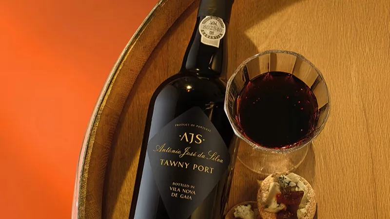 Bottle of AJS Tawny Port next to a glass of dark red wine and two small pastries on a wooden surface with an orange background.