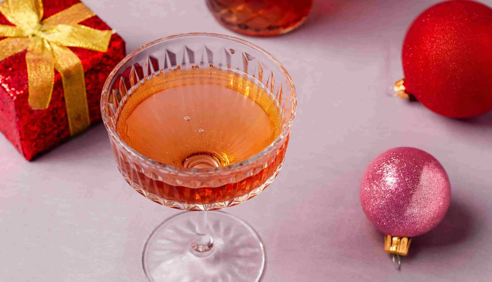Crystal coupe glass filled with amber-colored cocktail, surrounded by red and pink Christmas baubles and a red gift box with a gold ribbon on a light pink surface.
