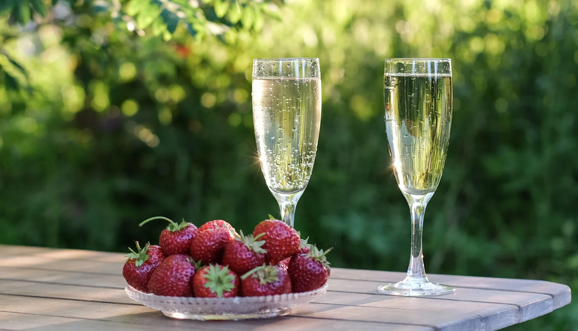 Two glasses of sparkling wine on a wooden table with a plate of fresh strawberries in an outdoor garden setting.