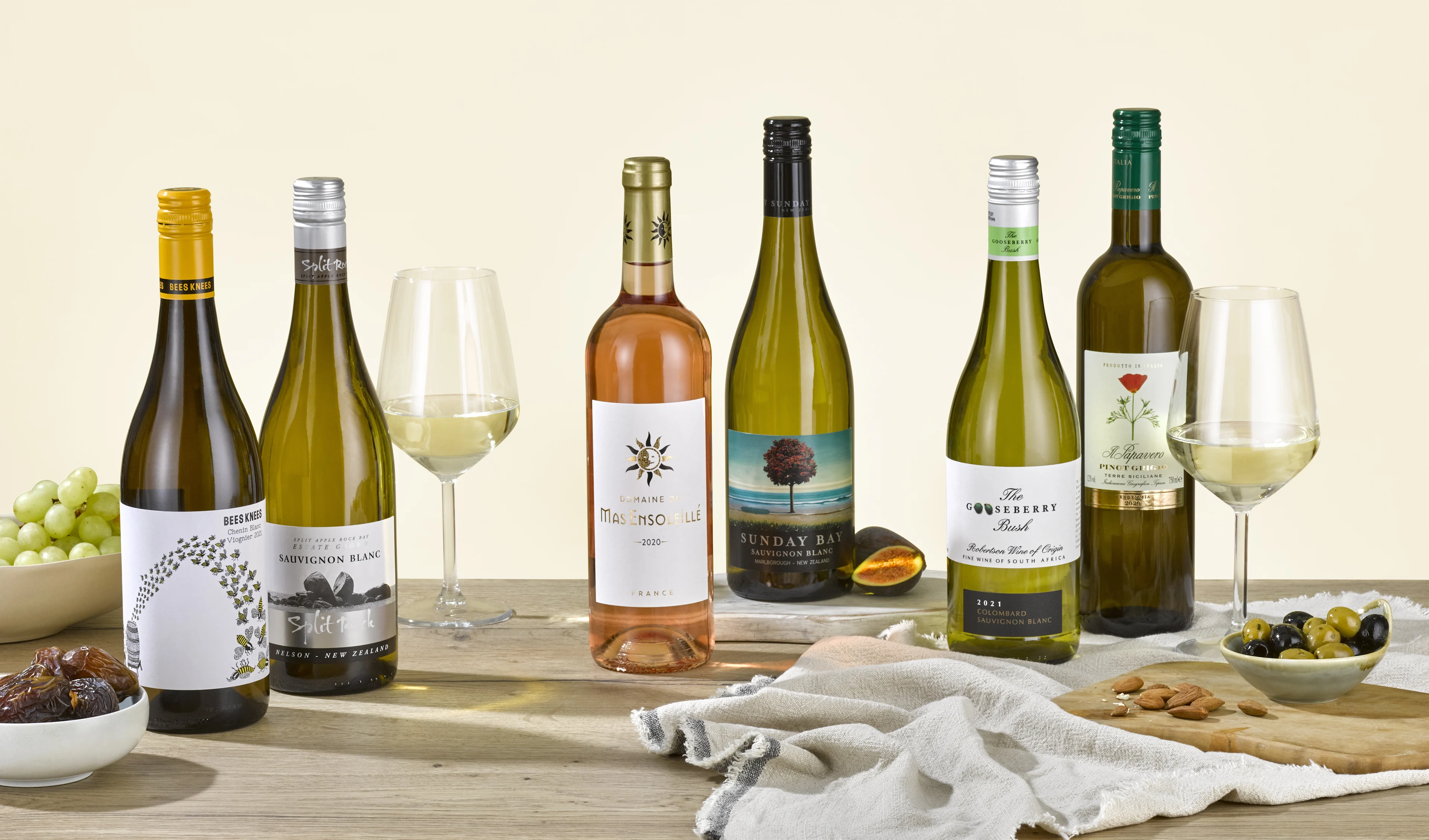 Selection of white wine bottles with filled wine glasses, grapes, olives, dates and almonds on a wooden table.