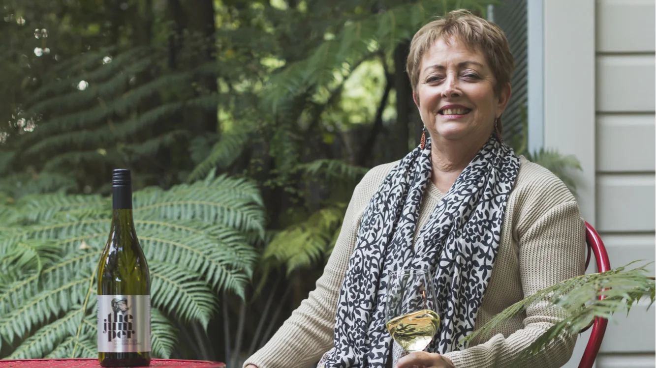 Person seated outdoors holding a glass of white wine beside a table with a wine bottle and a dog.